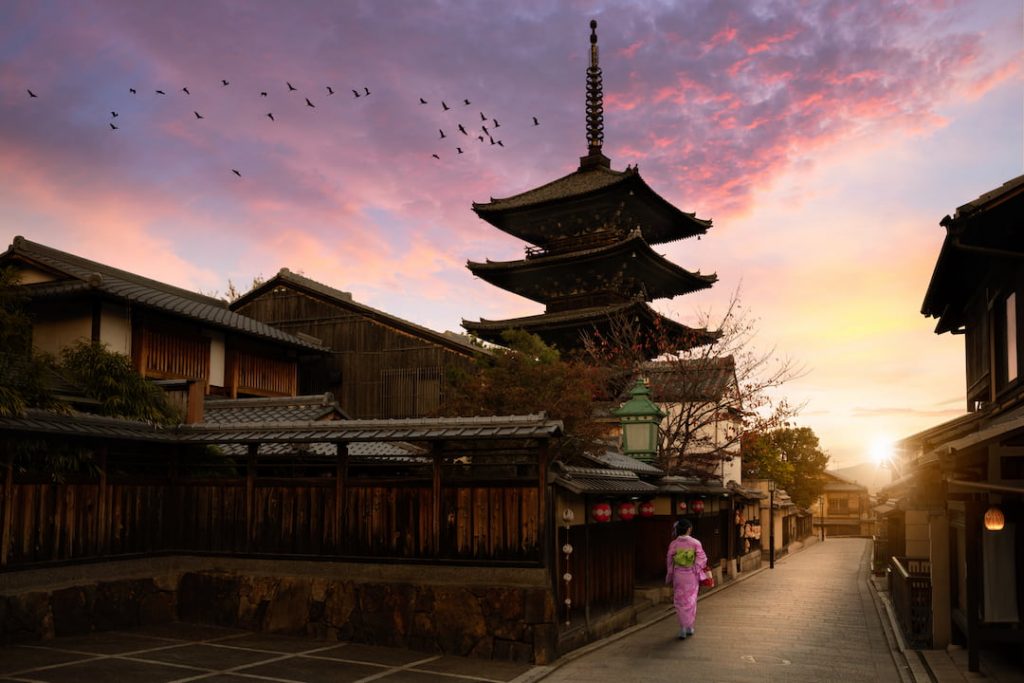 Traditional architecture in Japan, the sun has set, the sky is pink and birds fly across a cloud as a women walks through a pathway