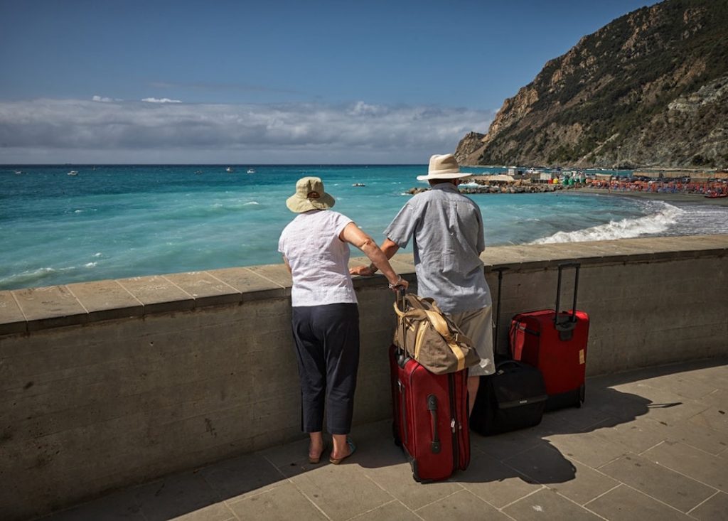 a man and woman stand looking over a bay with two red suitcases