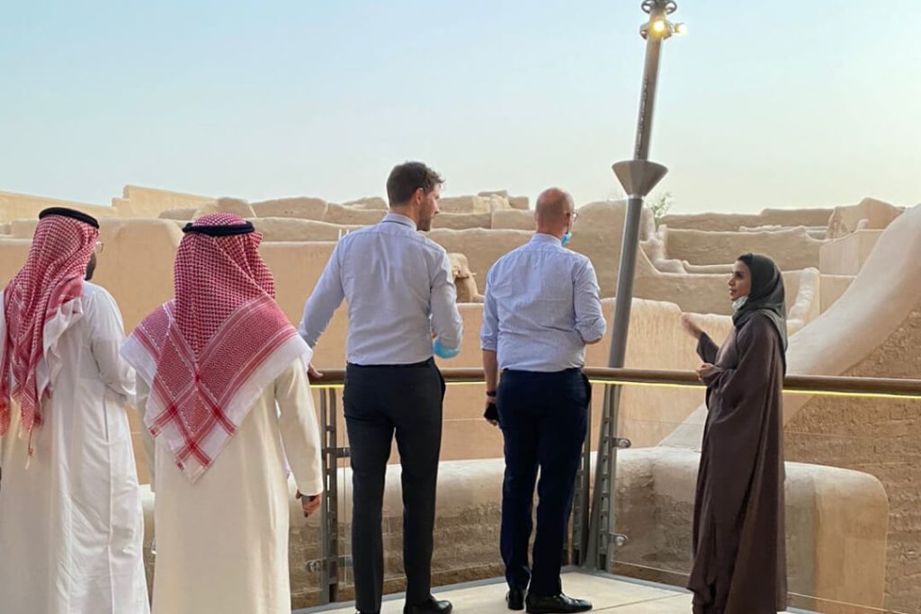 Four men and one woman in a mix of traditional Saudi clothing and business attire talk next to a ruin in Saudi Arabia.