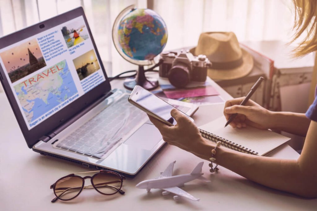 Tips for travelling to Saudi Arabia: a woman is sitting down at a table as she plans a trip using her smart phone, pen and paper, and computer.