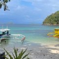 Two outrigger boats moored in turquoise water off a white-sand beach in the Philippines, with green islands on the horizon