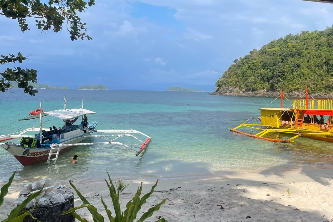 Two outrigger boats moored in turquoise water off a white-sand beach in the Philippines, with green islands on the horizon