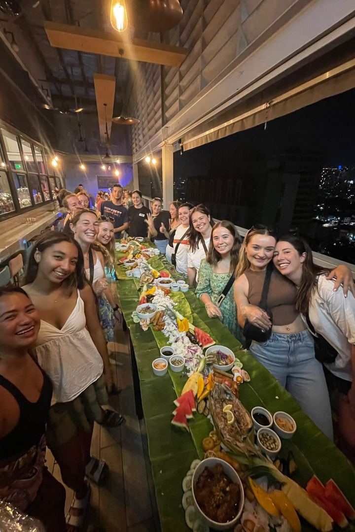 A group of travelers smiling around a long table covered in banana leaves and Filipino food, set up for a boodle fight