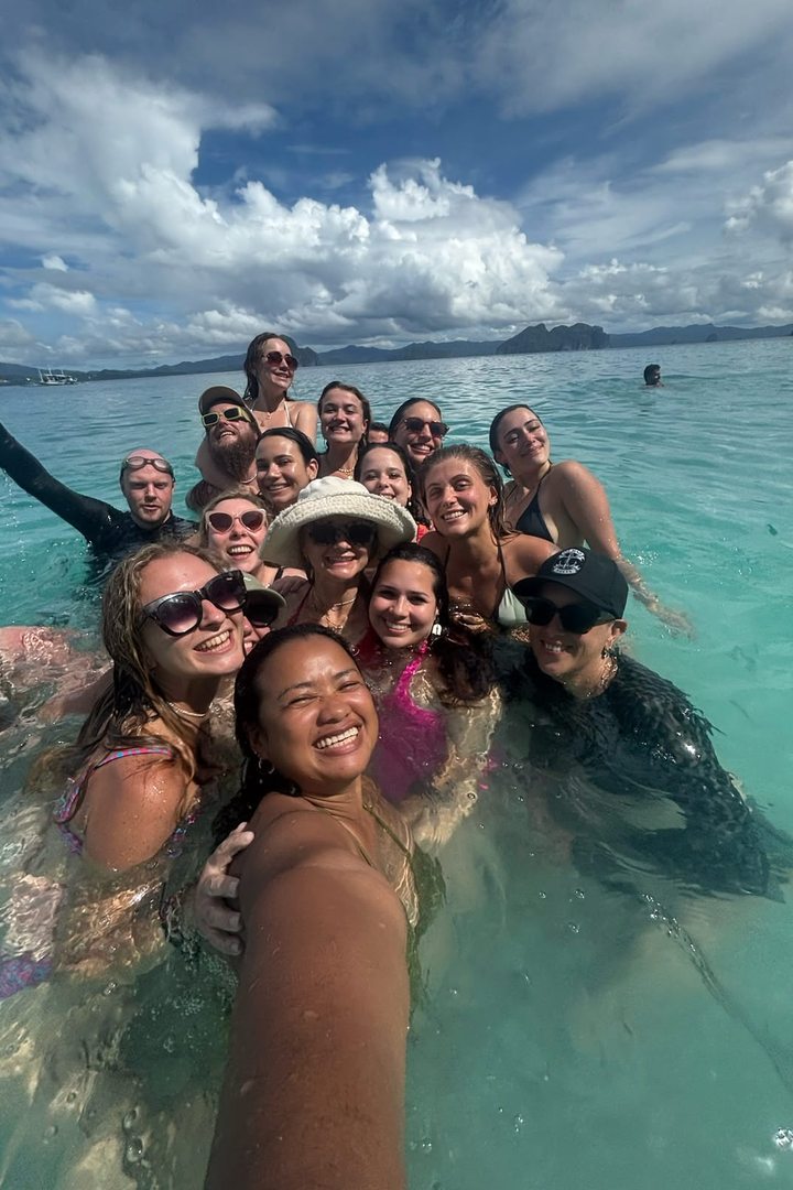 Diana Legaspi taking a selfie in turquoise water with a large group of smiling travelers from her tour