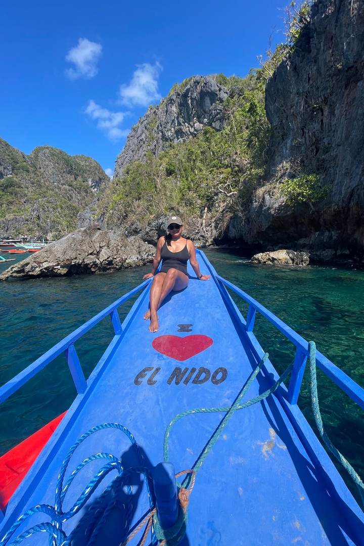 Diana Legaspi sitting at the bow of a blue outrigger boat painted with I love El Nido, sailing between limestone cliffs in Palawan