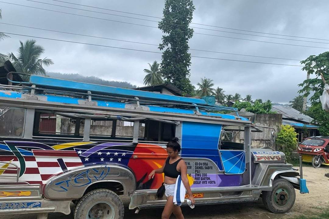 A colorfully painted jeepney parked under palm trees in the Philippines, with a traveler standing beside its open back door