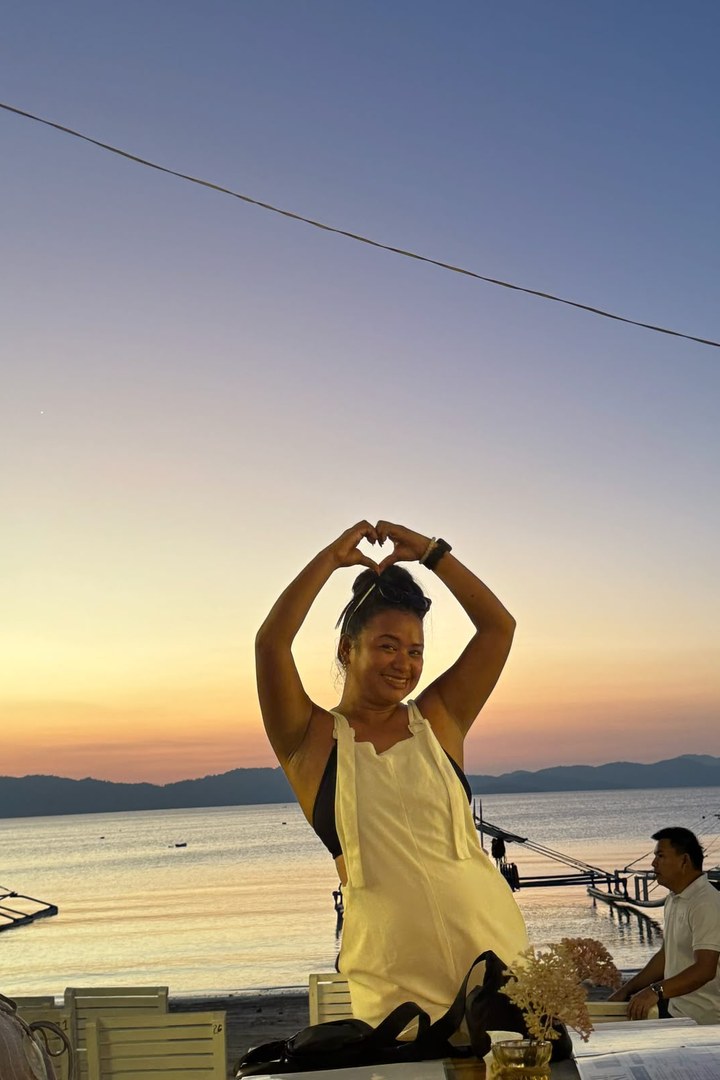 Tour guide Diana Legaspi making a heart shape with her arms on a beach at sunset in the Philippines, with outrigger boats in the background