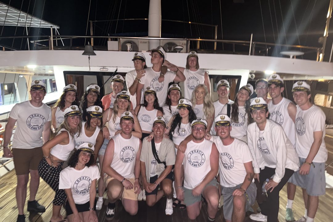 A large group of young travelers in matching white T-shirts and captain hats posing together on the deck of their boat at night in Croatia