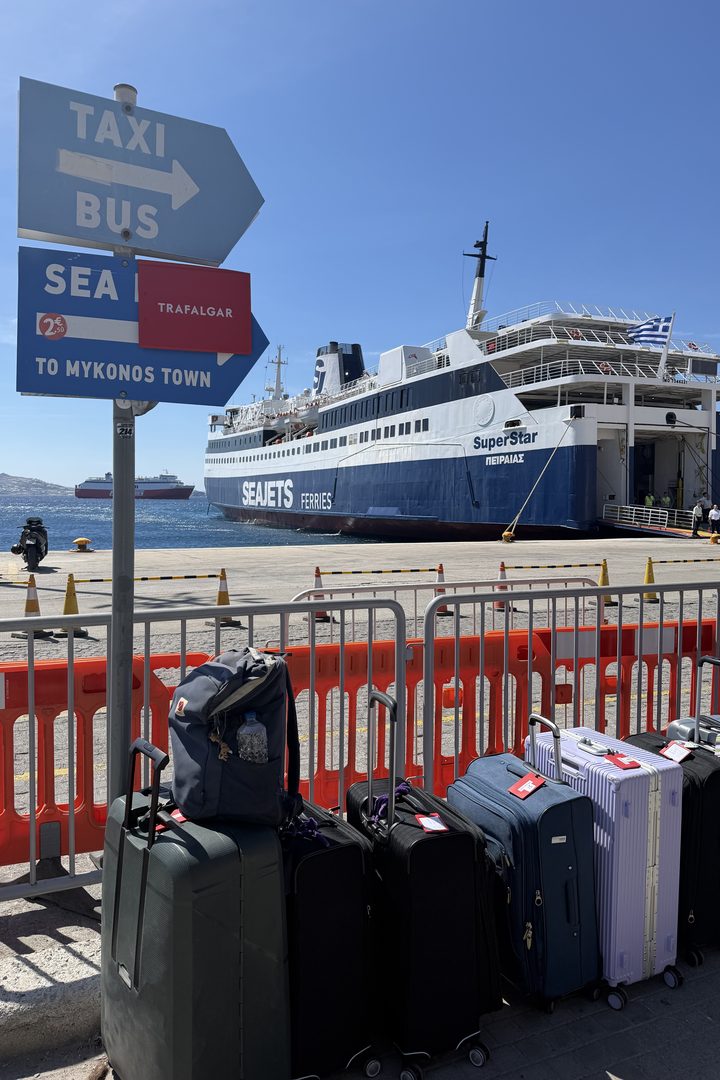 Suitcases lined up at Mykonos port next to a sign for taxis, buses and Mykonos Town, with a large passenger ferry docked behind