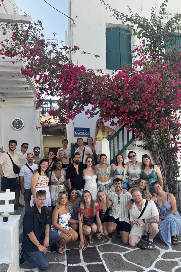 A group of travelers posing under pink bougainvillea in a whitewashed street in Mykonos Town, Greece