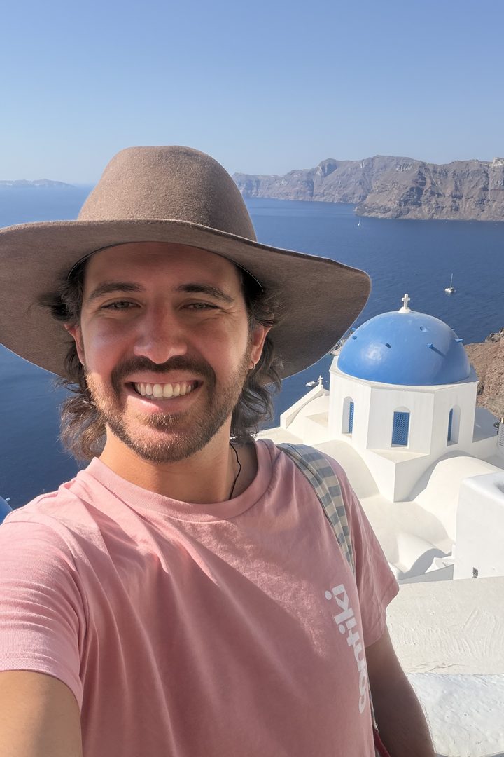 Trip manager Tom Whelan in a wide-brimmed cowboy hat taking a selfie above a blue-domed church in Oia, Santorini, with the caldera behind him